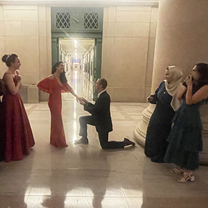 A photo of Kevin Greenman and Aline Vargas Manriquez at MIT's Lobby 7 with the infinite corridor behind them while Kevin is bent down on one knee holding Aline's hand