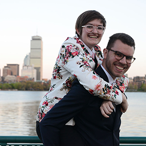 A photo of MIT alums Beau Rideout and Theresa Rideout with Theresa on Beau's back and the Charles River and Boston's Prudential Center in the background