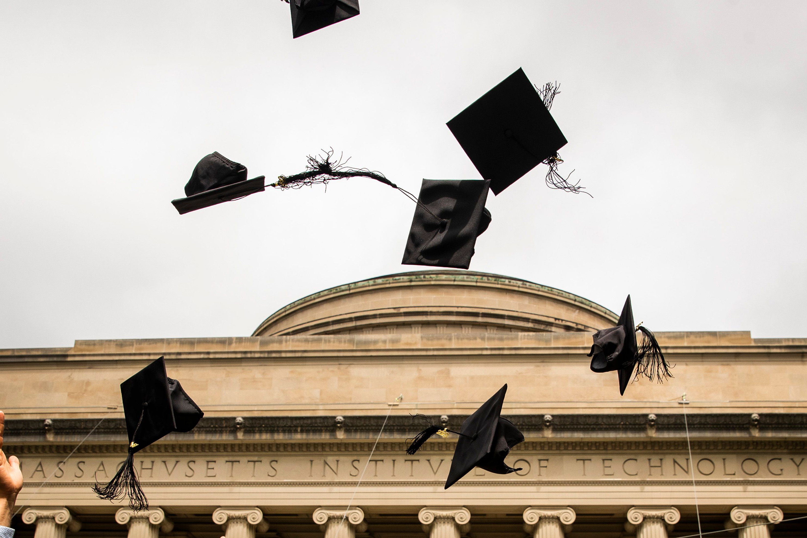 Graduation hats fly through the air against a background of gray sky and the top of the MIT dome