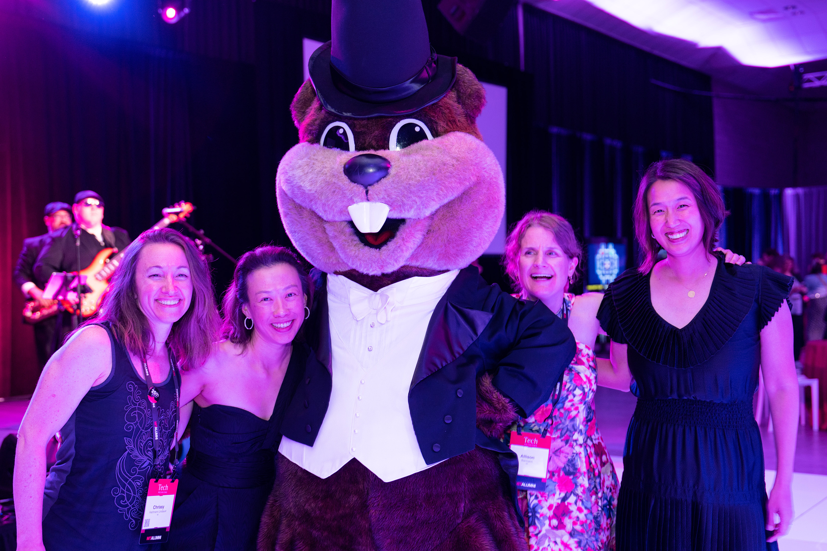 Four women in evening dress pose with Tim the Beaver dressed in a tux. Lighting is low and a band member can be seen in the background.