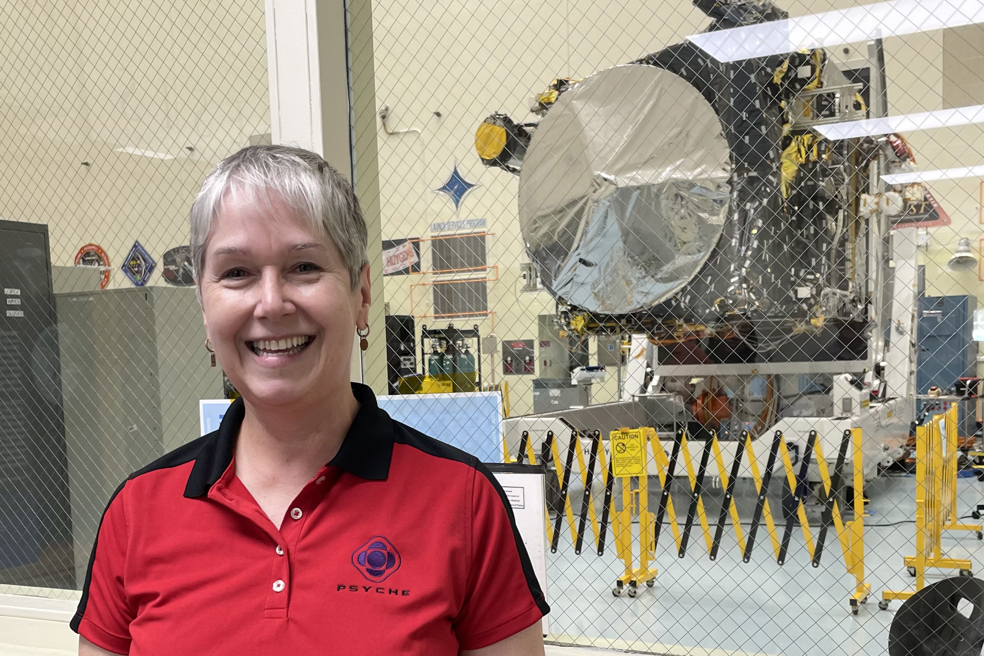 Lindy Elkins-Tanton poses in a red, collared Psyche shirt. A large silver disc on the spacecraft can be seen through the window behind her.