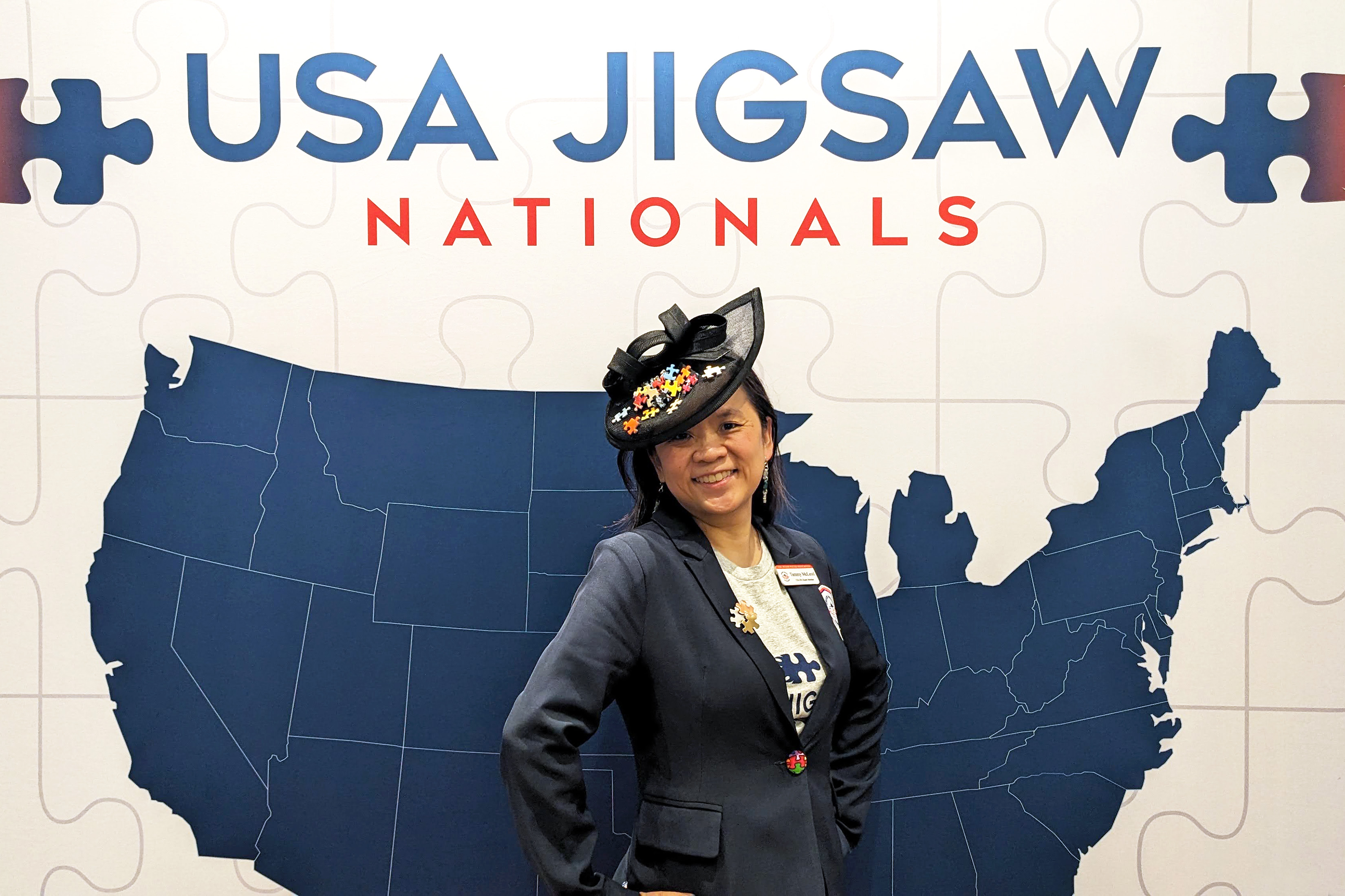 A photo of Tammy McLeod standing in front of a backdrop that says USA Jigsaw Nationals with a map of the US under the words