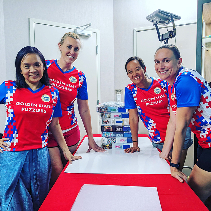 A photo of Tammy McLeod MIT alum with three other women wearing shirts that say Golden State Puzzlers with a stack of puzzles on a table