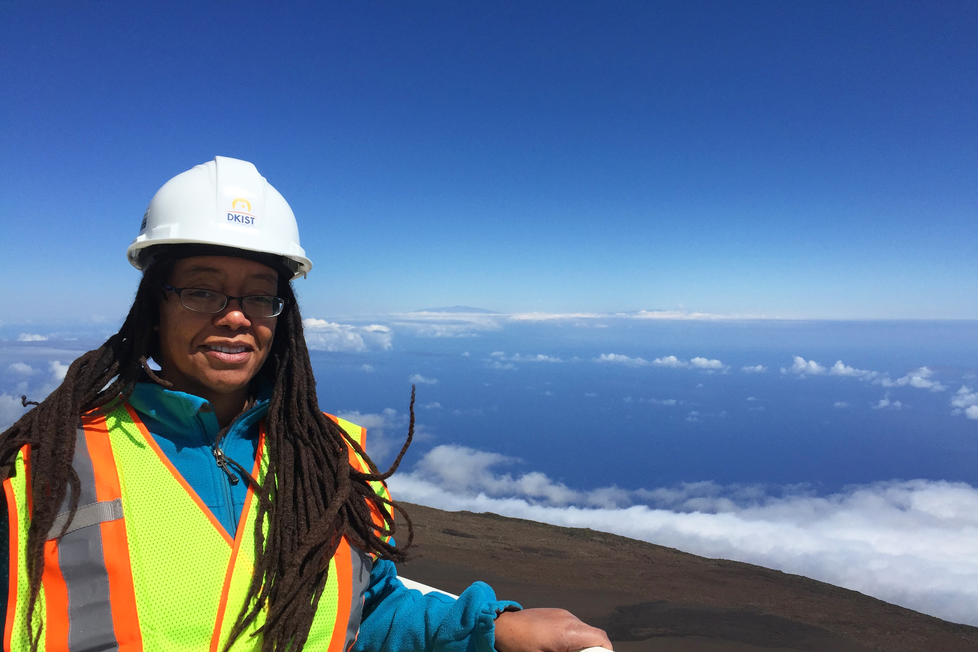 A photo of MIT alum Dara Norman wearing a hard had and a yellow reflective vest with her hand leaning on a railing and a blue sky and landscape behind her