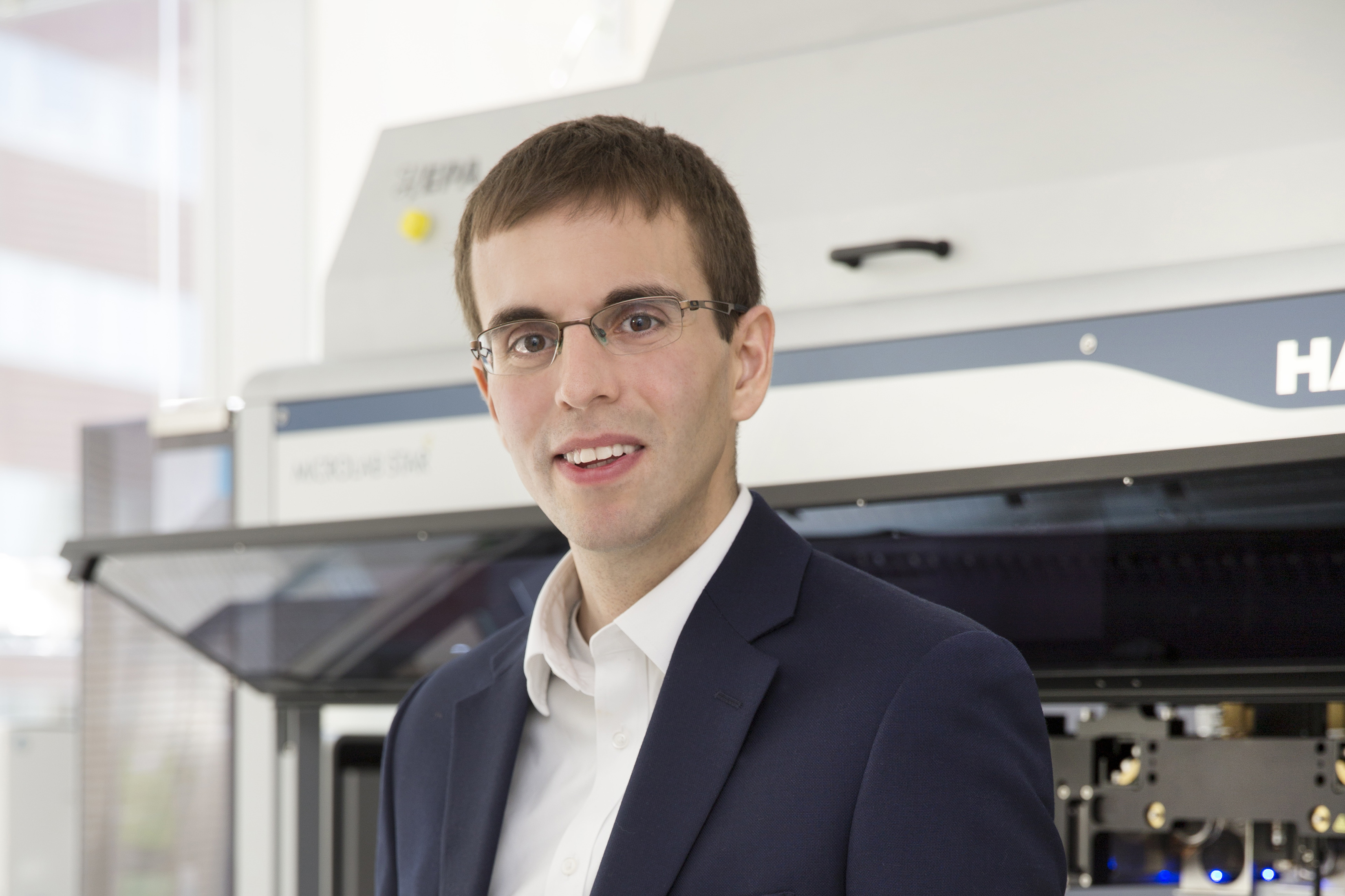 Viktor Adalsteinsson portrait. He is wearing a dark jacket and white shirt and some lab machinery is visible behind him.