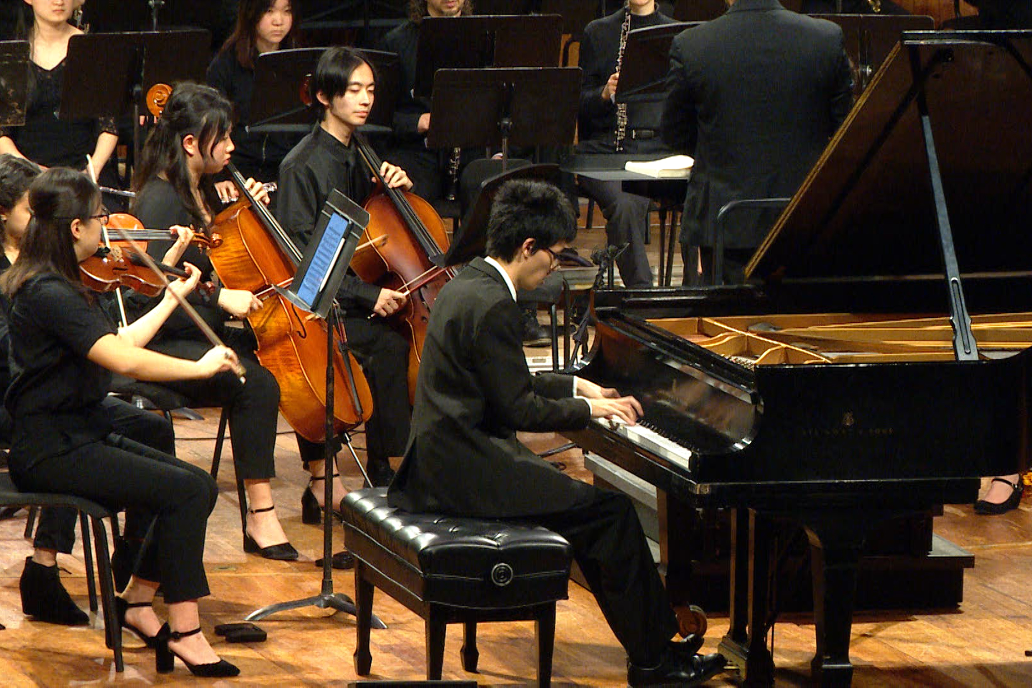 A pianist in a black suit sits on a bench playing a grand piano. Members of an orchestra appear behind him, notably two cellists and two violinists. Black music stands obscure other people.