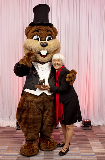 Sze-Wen Kuo is shown hugging Tim the Beaver and holding a small beaver statue. Tim is wearing a tuxedo.