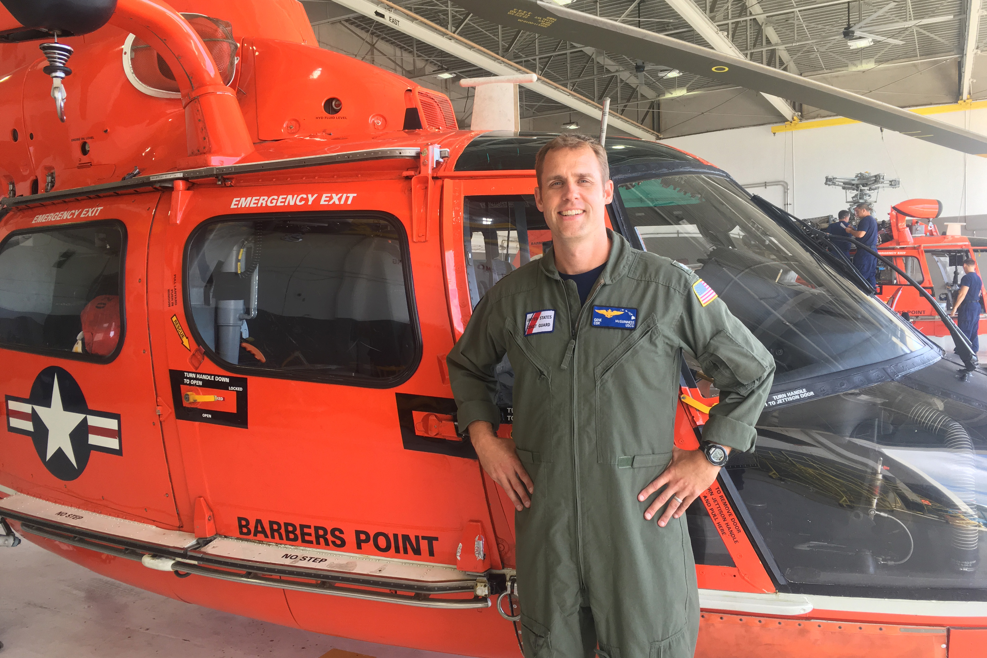 Gene McGuinness stands in front of a red helicopter wearing an olive flightsuit with a Coast Guard patch on it.