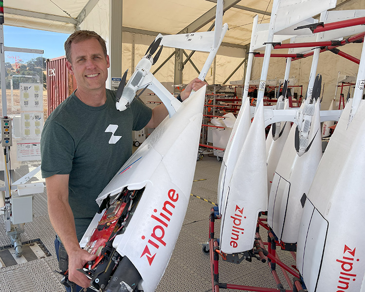 Eugene McGuinness is shown holding a Zipline delivery drone, which is nearly as big as he is. Several more are arrayed at right.