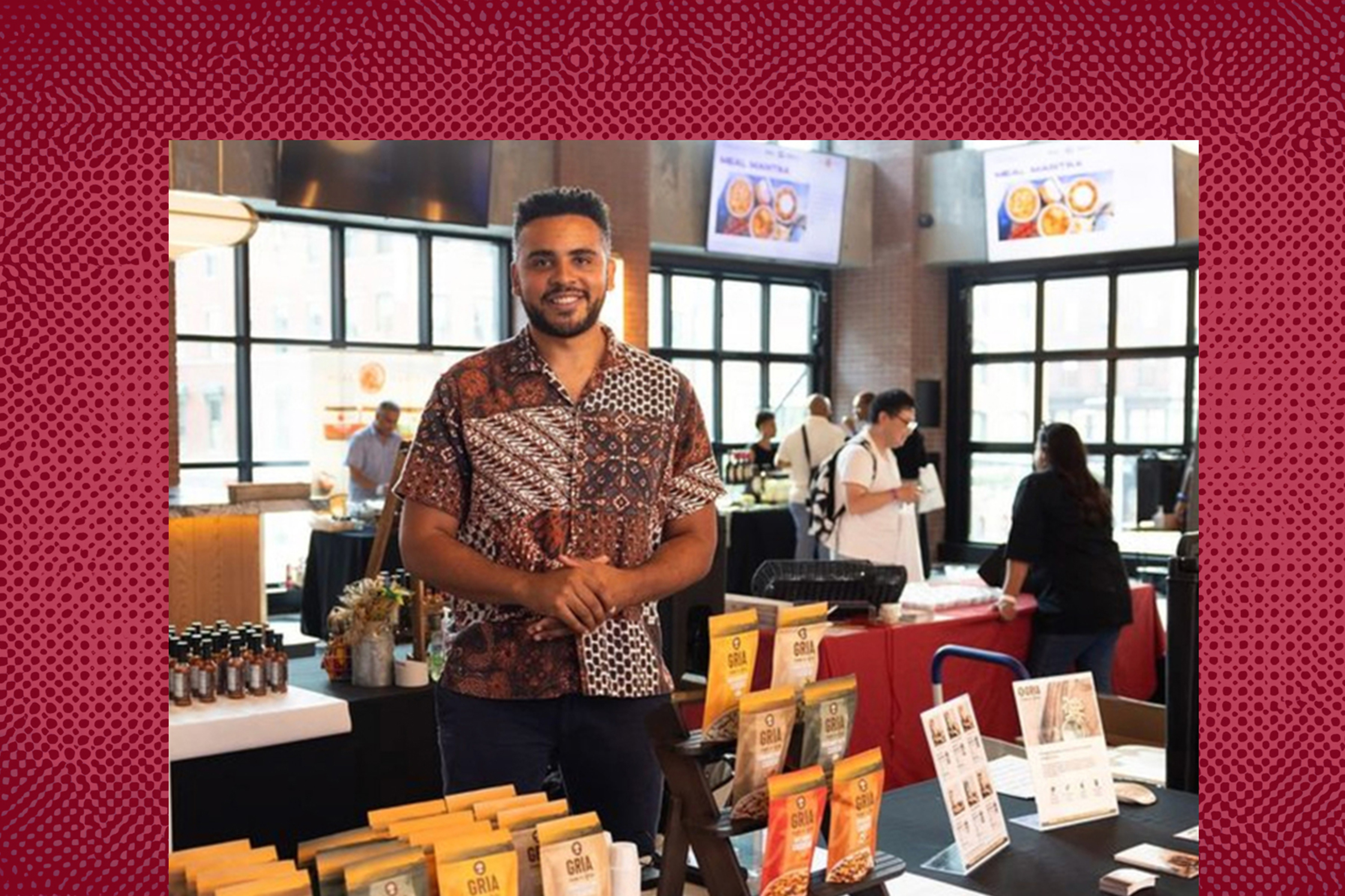 Joshua Reed-Diawuoh stands behind a table featuring GRIA products and small cups. Other tables and shoppers are visible in the background. The image has a red patterned border.