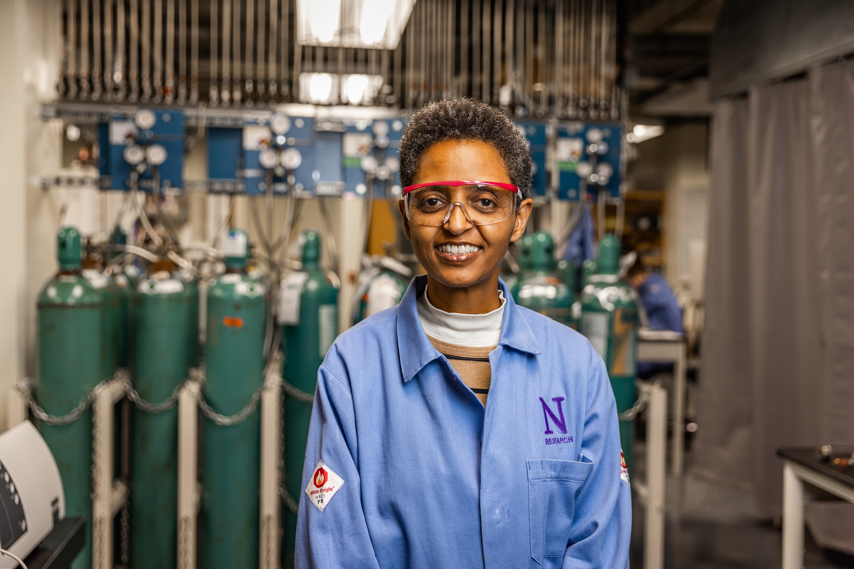 Sossina Haile portrait. She is wearing a blue lab coat featuring a purple N. She is standing in front of an array of narrow green tanks and other equipment.