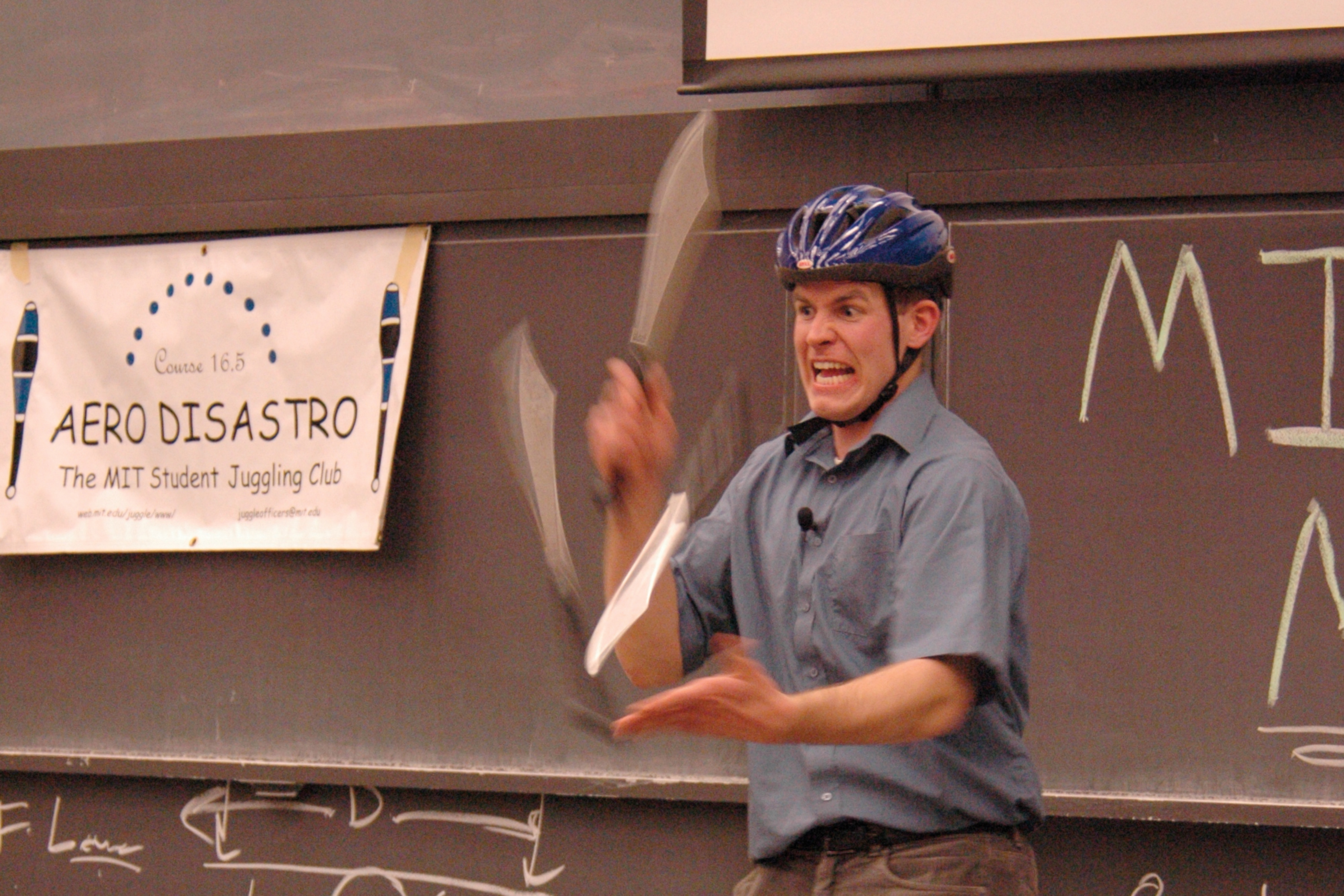 A photo of David Rush in a classroom with a blackboard behind him wearing a helmet and juggling knives