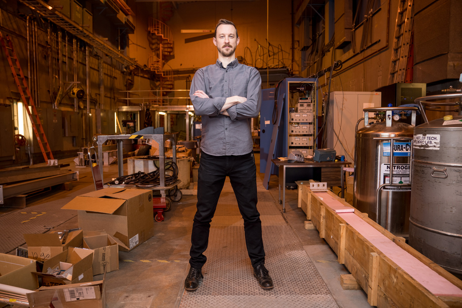 Zachary Hartwig stands arms crossed in what looks like a large workshop. Nitrogen tanks, boxes, ladders, wires, and other materials can be seen in the background.