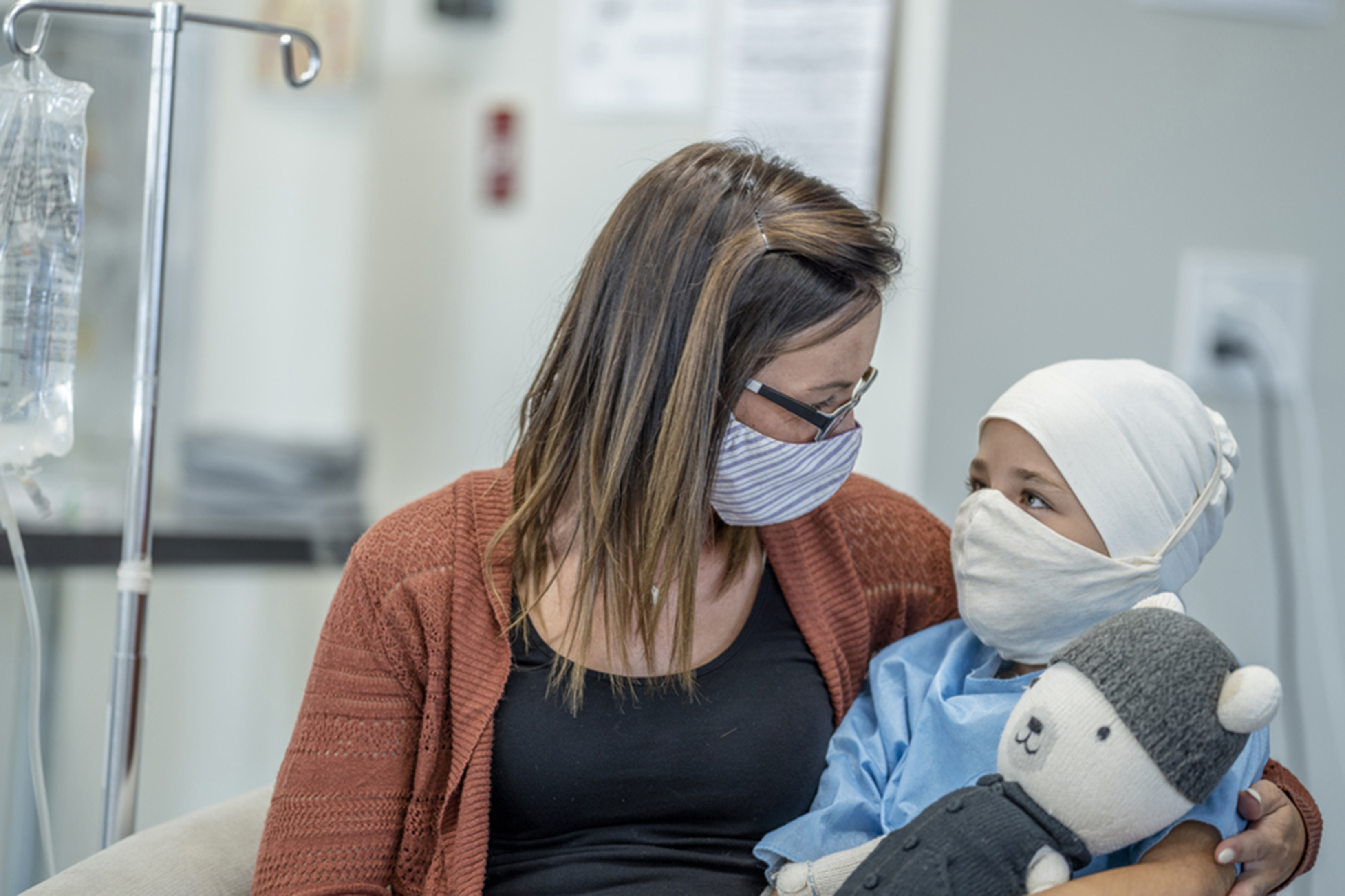 A mother hugs her young daughter while she receives cancer treatment. Both are masked, and the child holds a plush toy