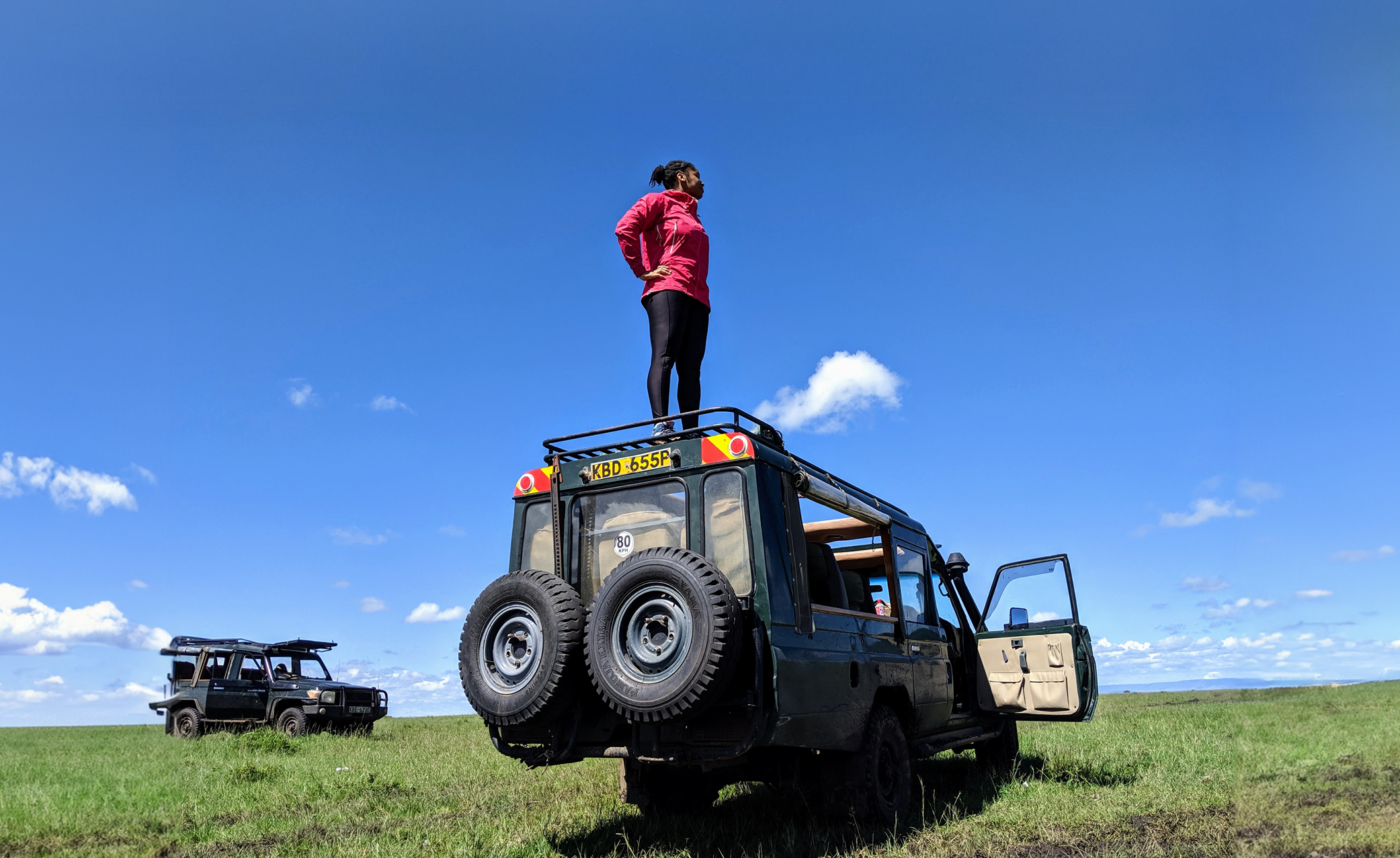 Jamila Smith-Dell Stands on the top of a Land Rover on a safari, looking off to the right with blue sky and grass below