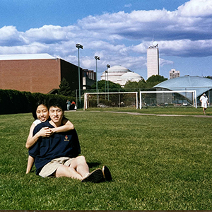 A photo of Benson Yang and Carina Yang sitting in a grass field at MIT with Kresge auditorium and the great dome in the background