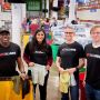 Four MIT Alumni wearing black shirts with MIT Alumni on the front stand in a warehouse holding clothes